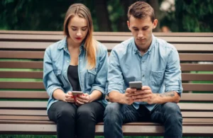 Dos personas sentadas en un banco, absortas en sus teléfonos móviles, simbolizando la adicción a la tecnología, representando la necesidad de tratamiento con un psicólogo online en Ávila.