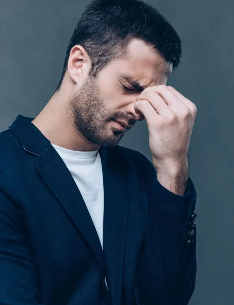 Hombre joven con signos de estrés y agotamiento, tocándose el puente de la nariz en señal de frustración. Ideal para ilustrar la importancia de buscar ayuda de un psicólogo online para el tratamiento del estrés. Terapia en línea para la gestión del estrés, disponible en cualquier momento y lugar.